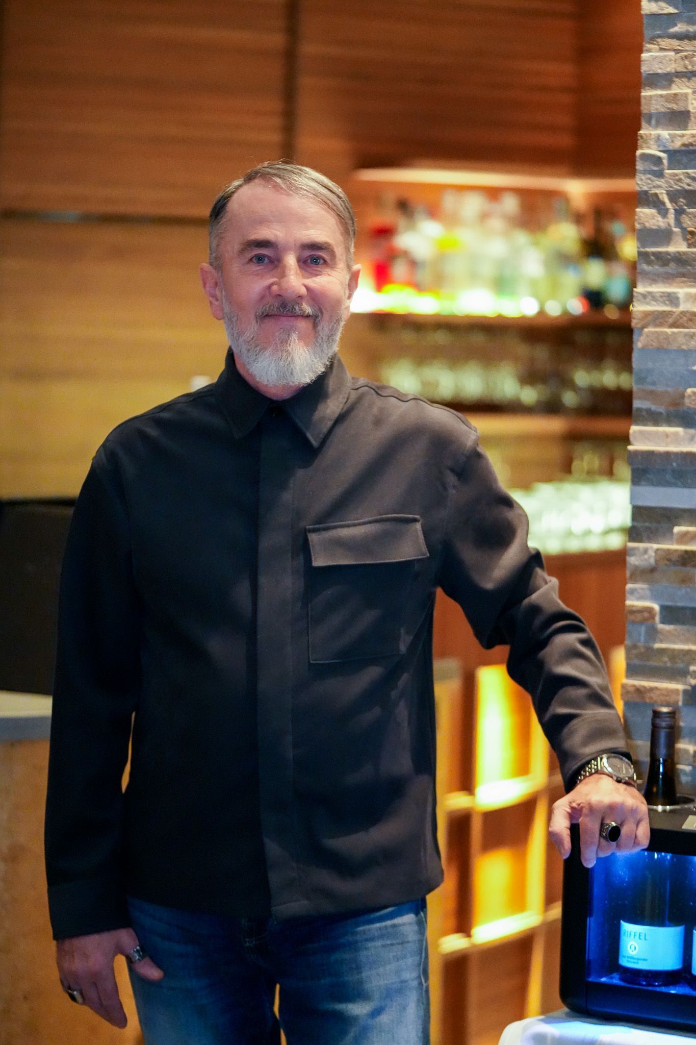 Ein Mann mit Bart und schwarzem Hemd steht in einer Bar neben einem Kühlschrank mit Weinflaschen.