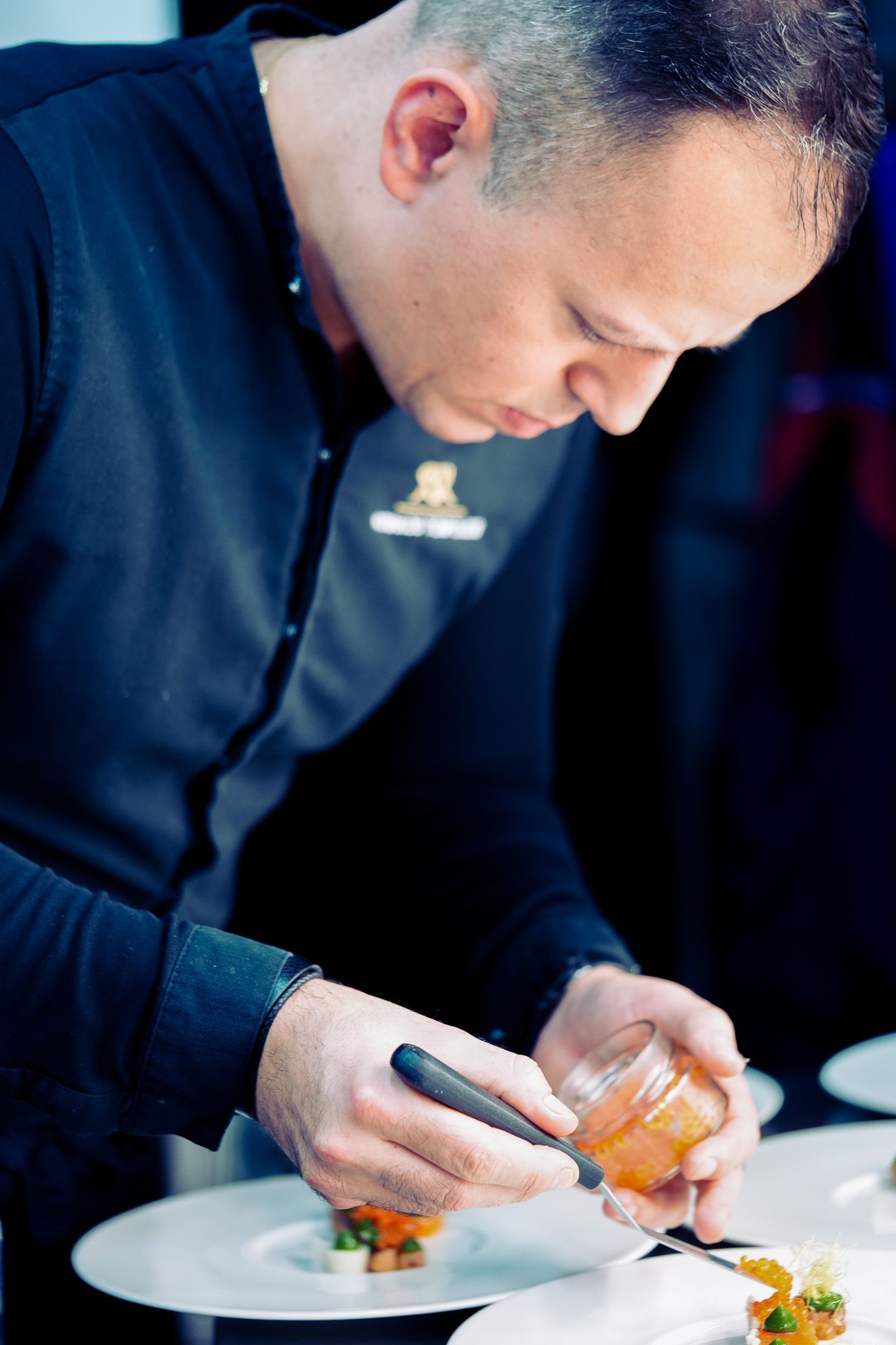 Ein Koch gibt Toppings auf ein Gericht auf einem weißen Teller. Dunkle Uniform, konzentrierter Gesichtsausdruck, drinnen.