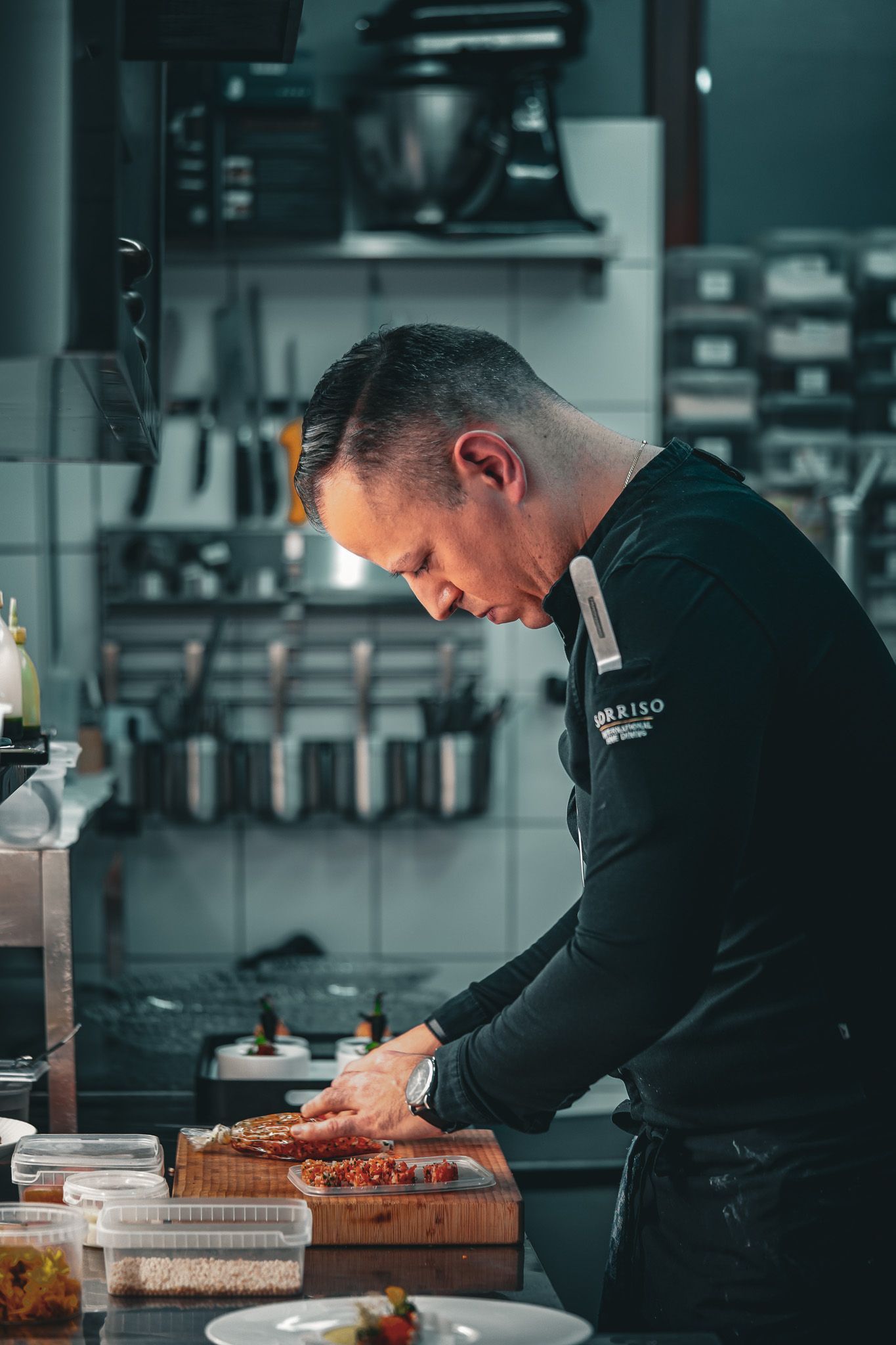 Ein Koch in schwarzer Uniform bereitet in einer Küche Speisen auf einem Holzschneidebrett zu.