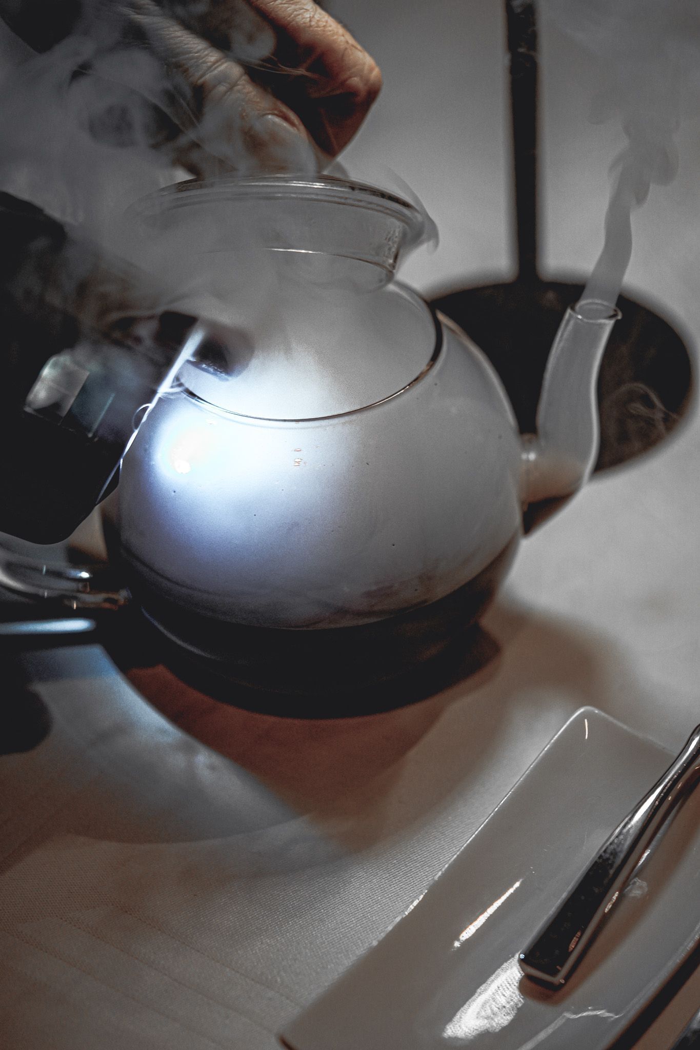 Eine Person gießt Dampf aus einer gläsernen Teekanne auf einen Tisch. Dampf steigt um die Kanne herum, in der Nähe des Silberbestecks, auf.