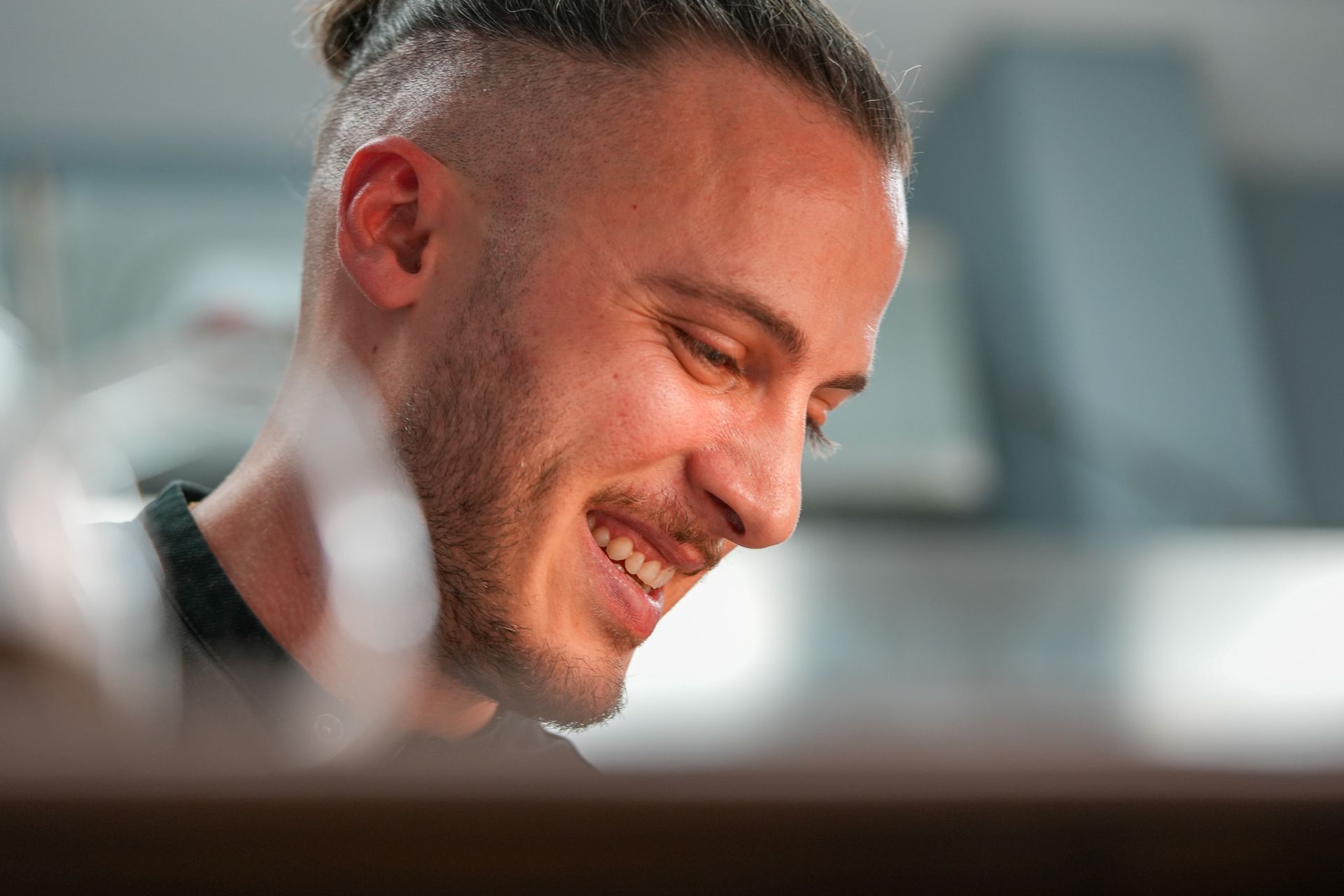 Mann mit hellbraunem Haar, lächelnd, den Blick gesenkt; verschwommener Küchenhintergrund.