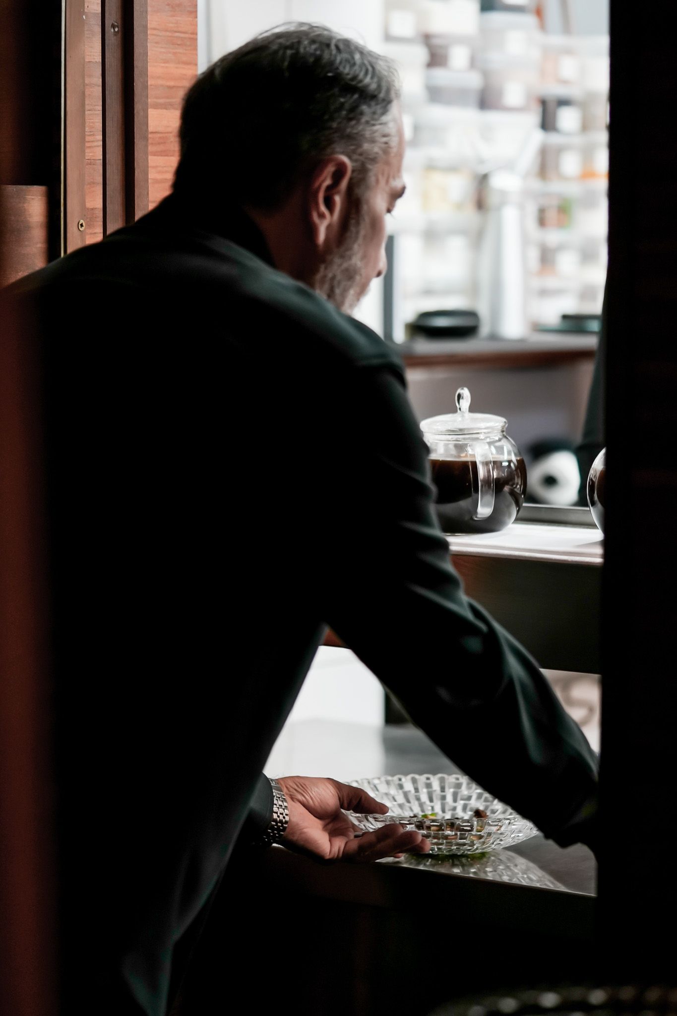 Ein Mann in einem dunklen Anzug beugt sich zu einer Theke in der Nähe eines Fensters. Er hält einen Teller in der Hand.
