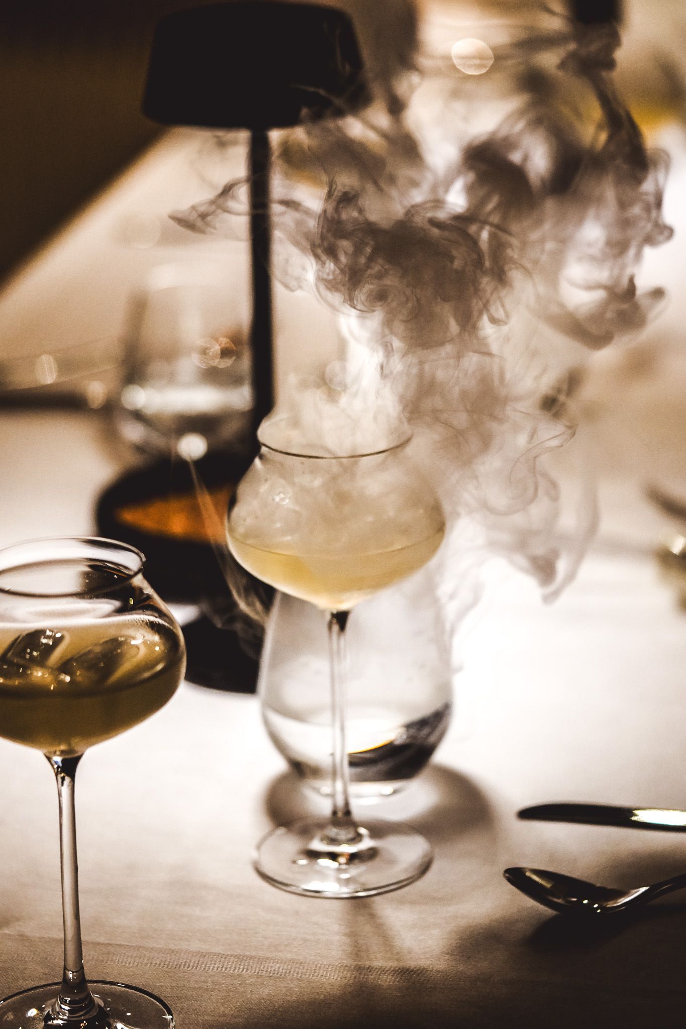 Ein Cocktailglas, das auf einem Tisch Rauch ausstößt, ein weiteres Glas im Fokus. Schwaches Licht.