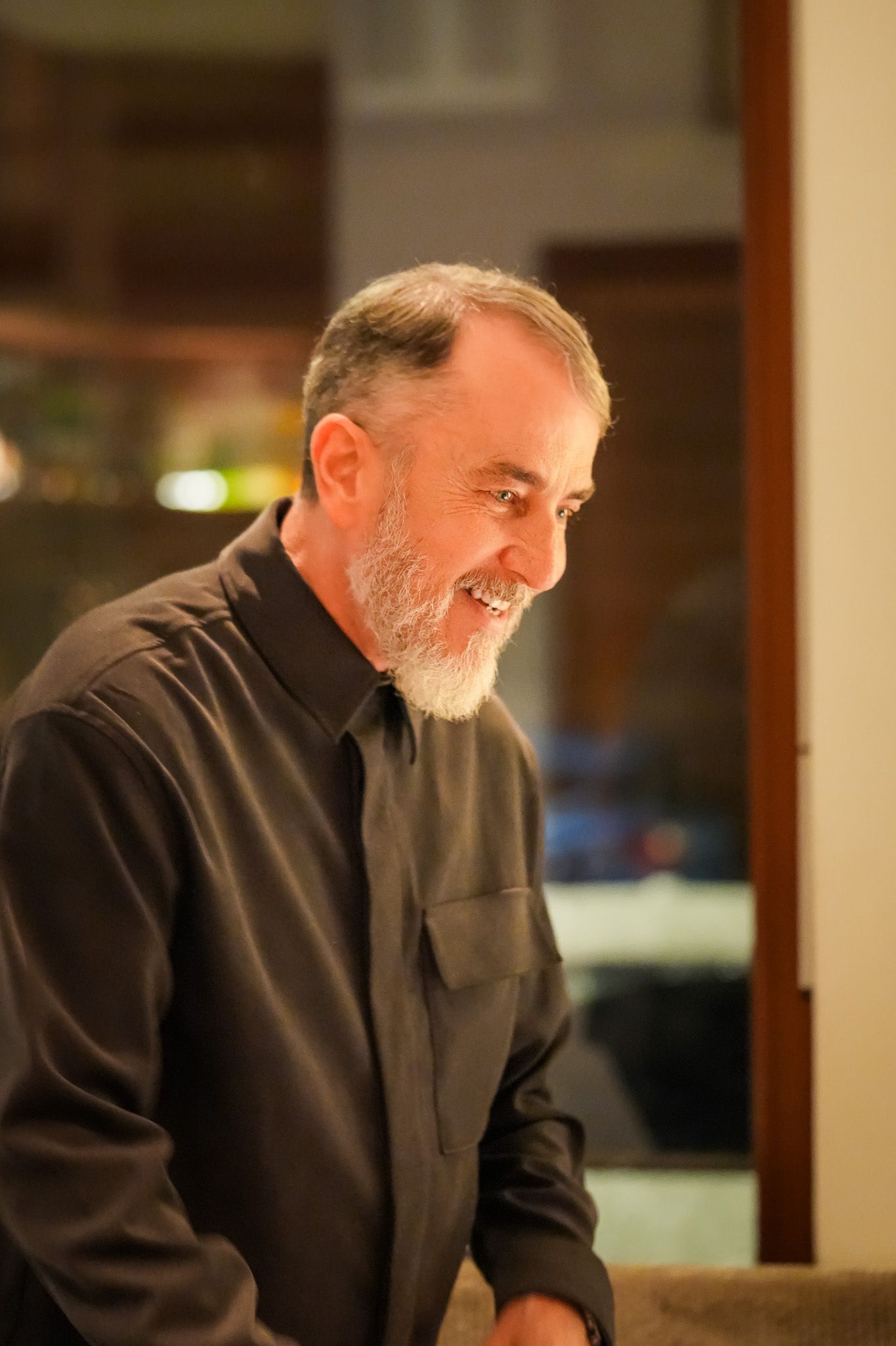 Ein Mann mit ergrauendem Bart lächelt in einem schwach beleuchteten Raum; er trägt ein dunkles Hemd mit Knöpfen.