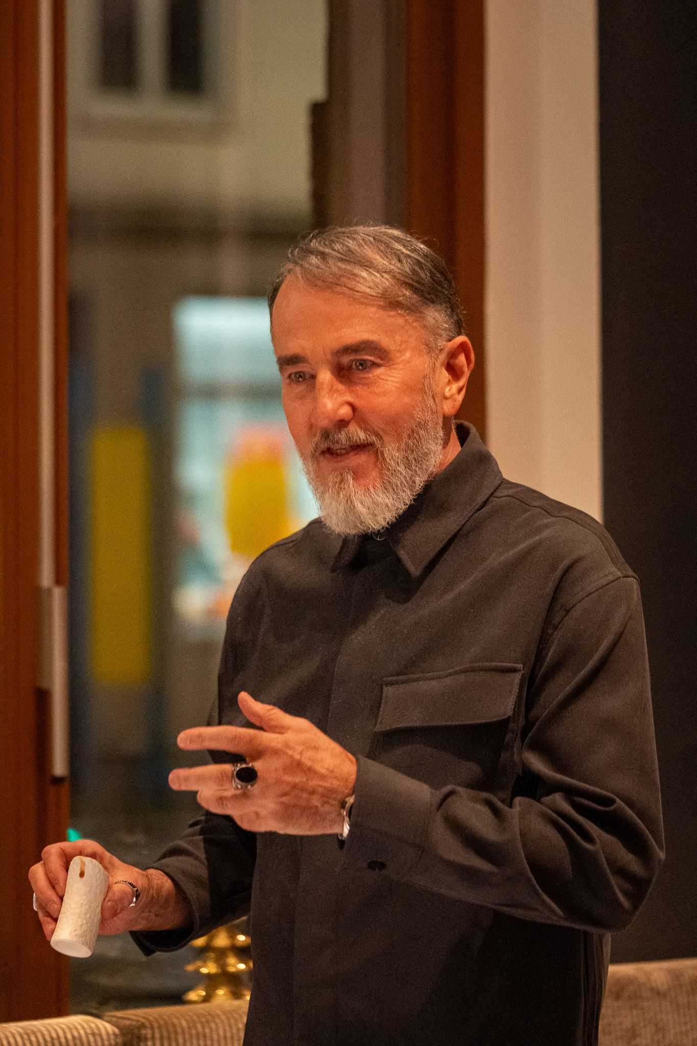 Ein Mann mit Bart gestikuliert, während er einen Gegenstand in der Hand hält und in einem Innenraum spricht.