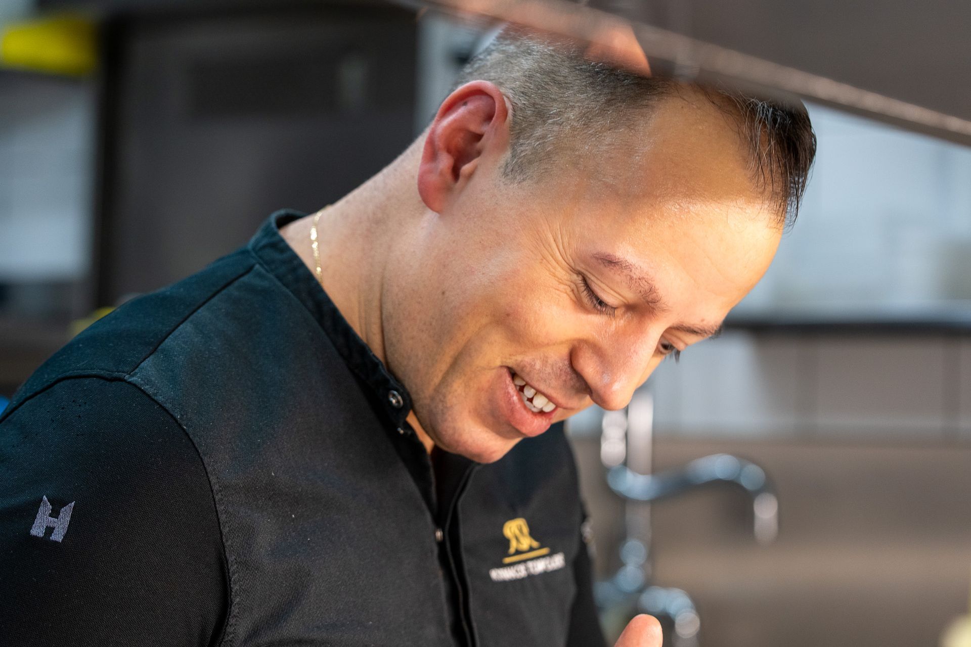 Ein Koch lächelt bei der Arbeit in der Küche; er trägt ein schwarzes Hemd mit Logo.