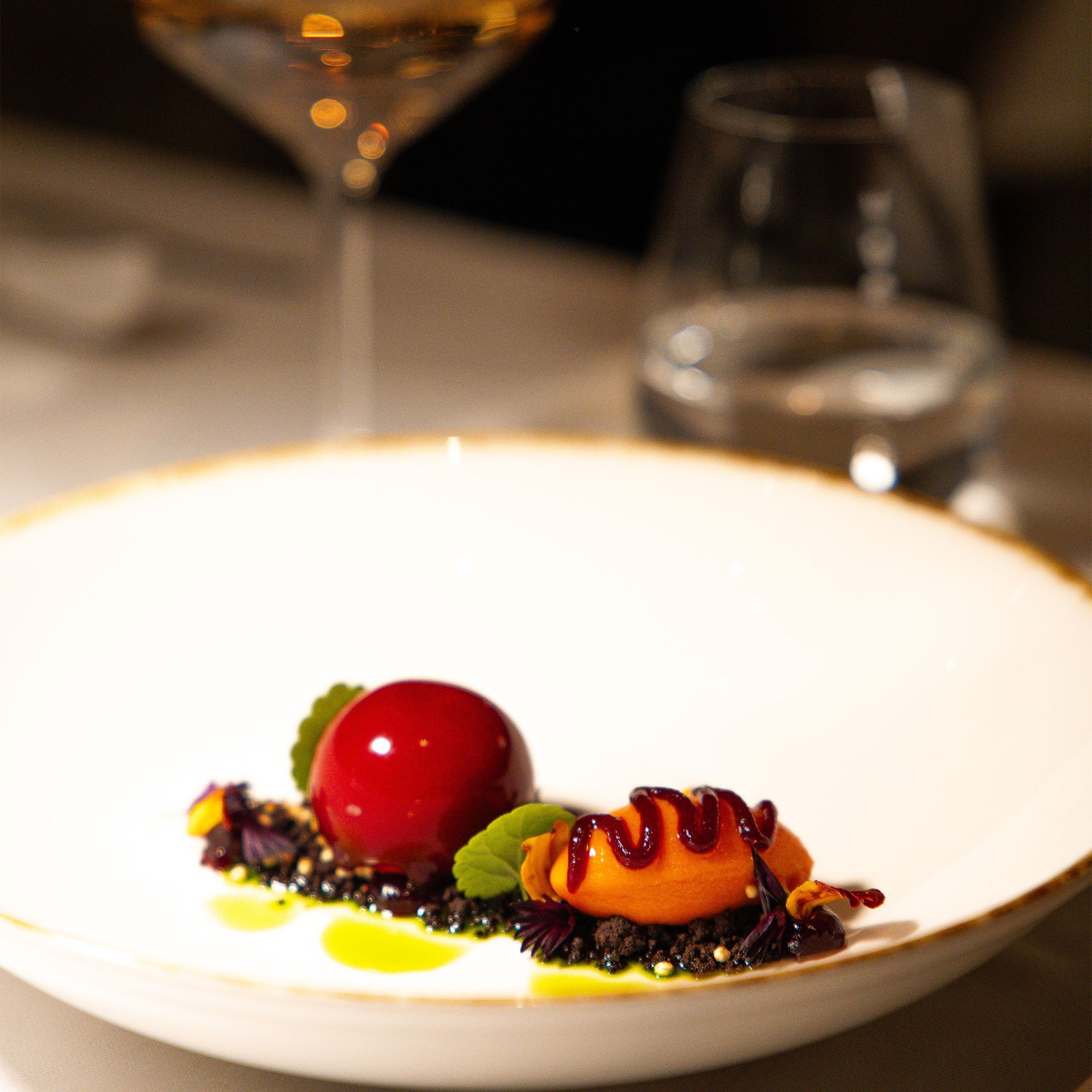 Ein Teller mit einem roten, kugelförmigen Dessert, einem Klecks Orange und schwarzen Streuseln, im Hintergrund ein Weinglas.