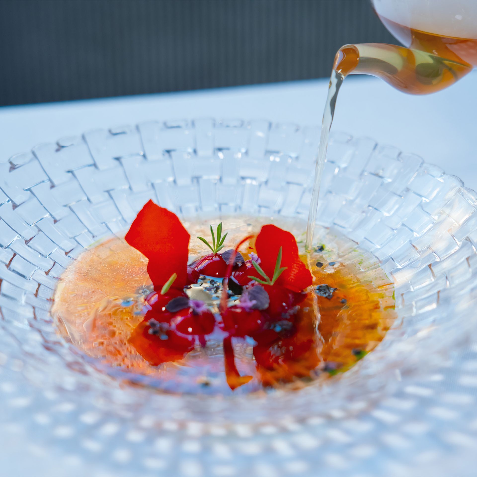 Eine durchsichtige Glasschüssel mit Lachs, Erdbeeren und einer klaren Flüssigkeit, die aus einer Teekanne gegossen wird.