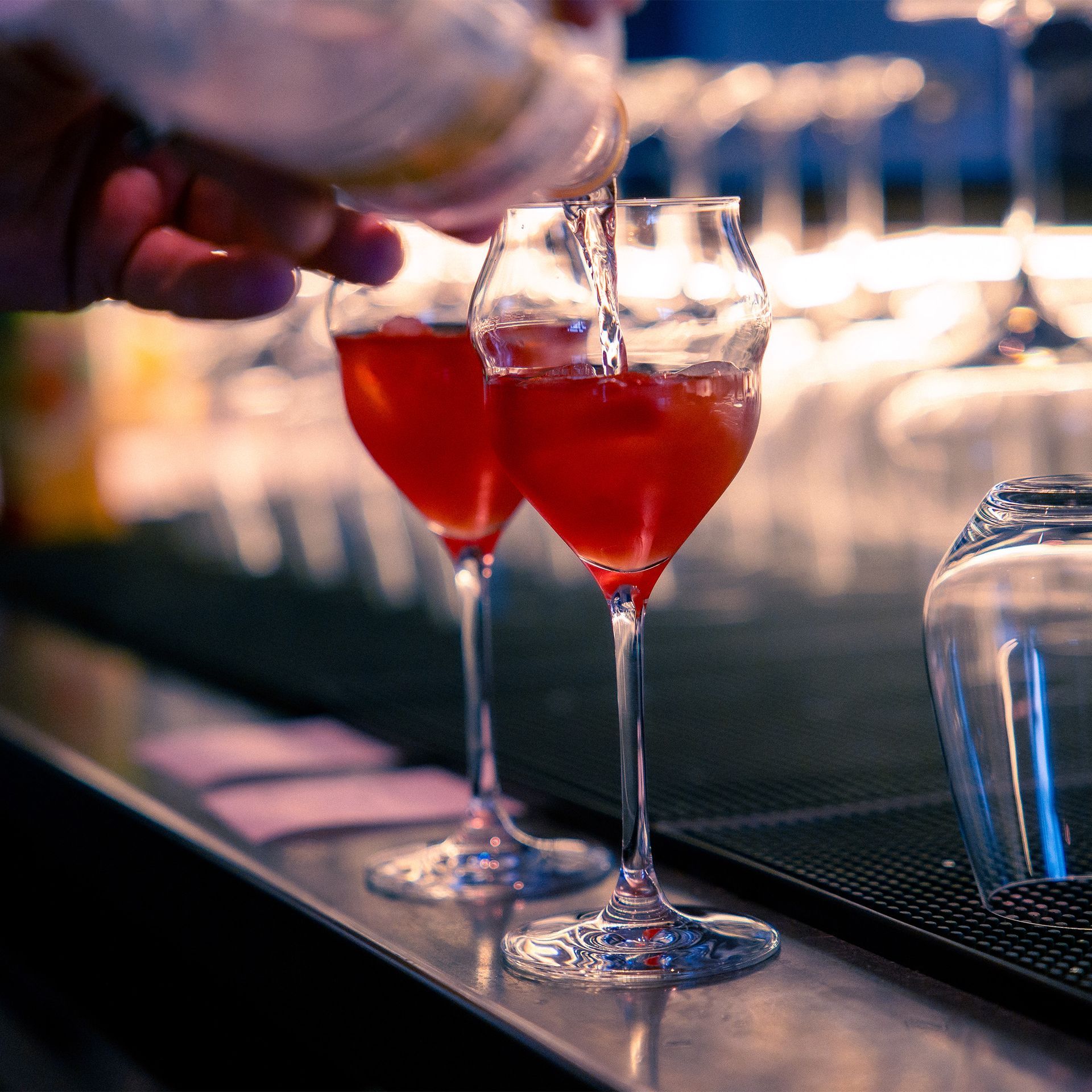 Ein Barkeeper gießt eine rote Flüssigkeit in ein tulpenförmiges Glas; daneben steht ein weiteres, ähnliches Glas auf der Bar.