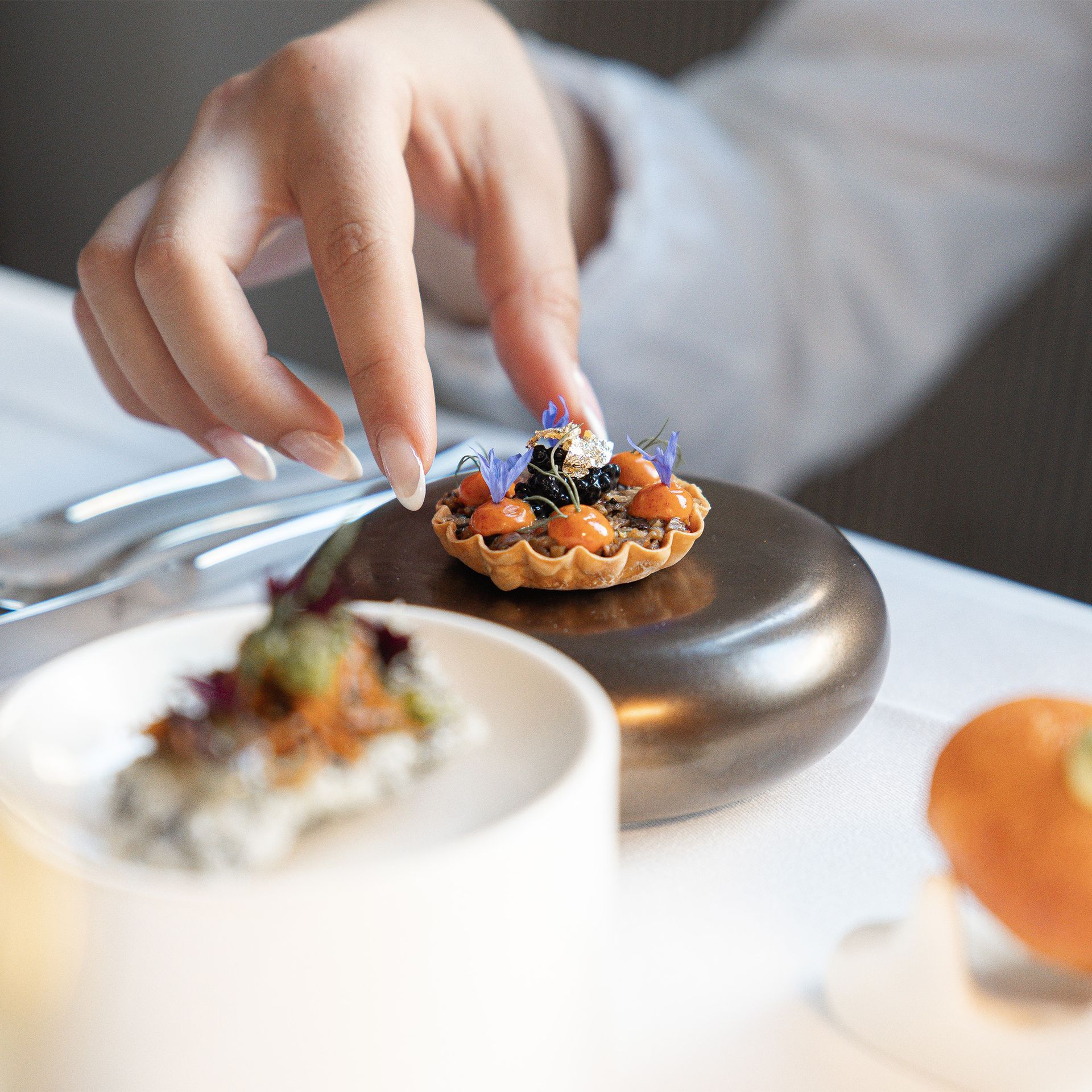 Eine Hand legt behutsam eine Garnierung auf ein Miniaturtörtchen. Szene in einem eleganten Speisesaal mit weißer Tischdecke.