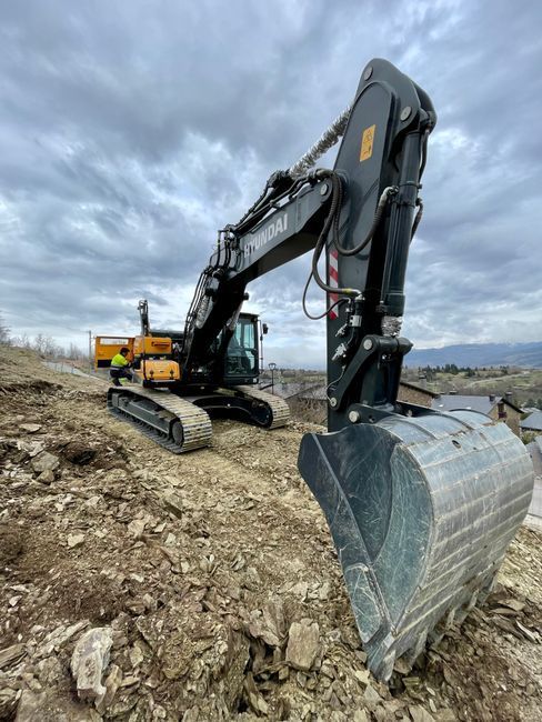 Excavator on a rocky construction site under a cloudy sky, with its bucket in the foreground