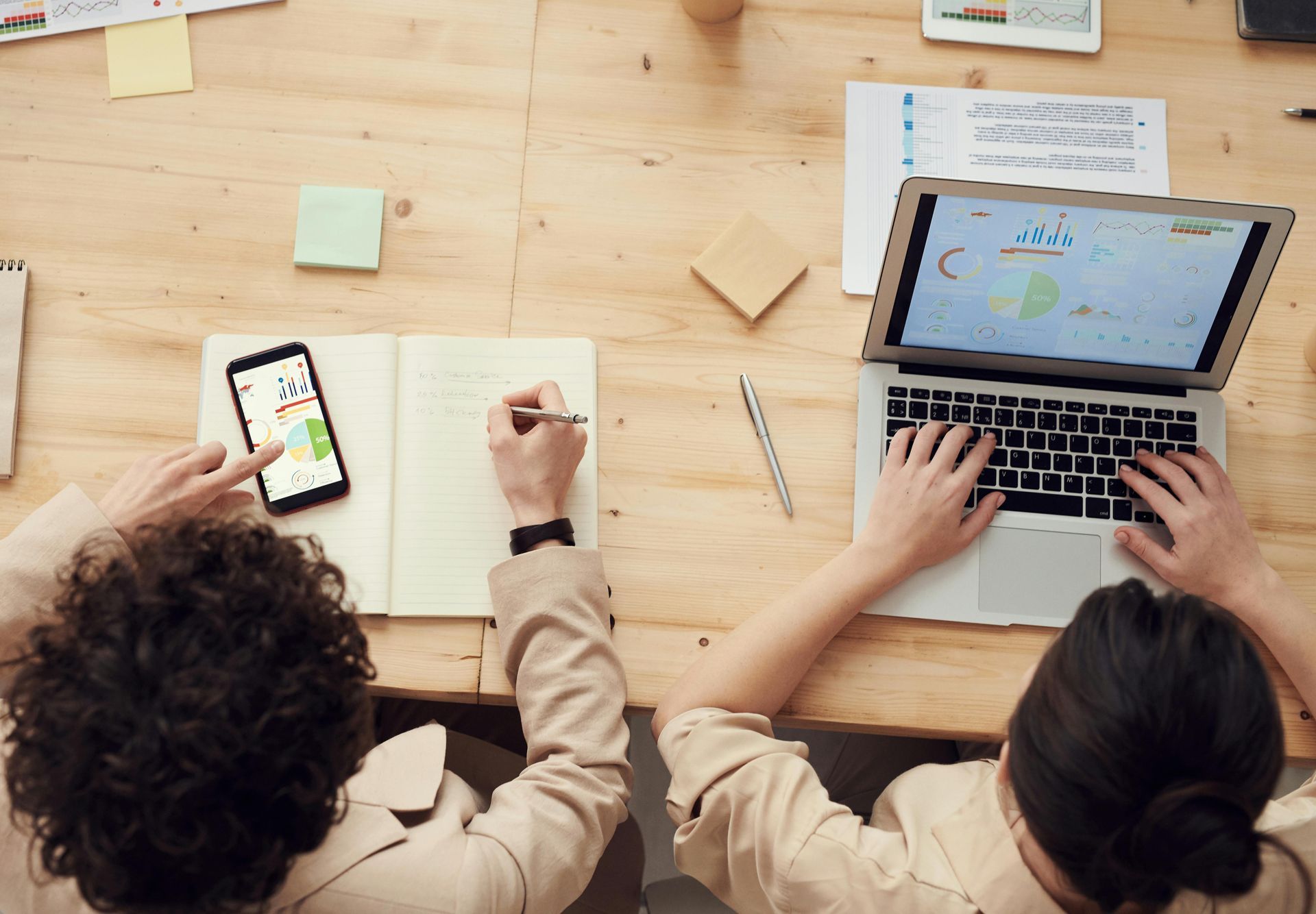 Two people collaborate at a wooden desk, using a laptop and smartphone with papers and notebooks nearby.