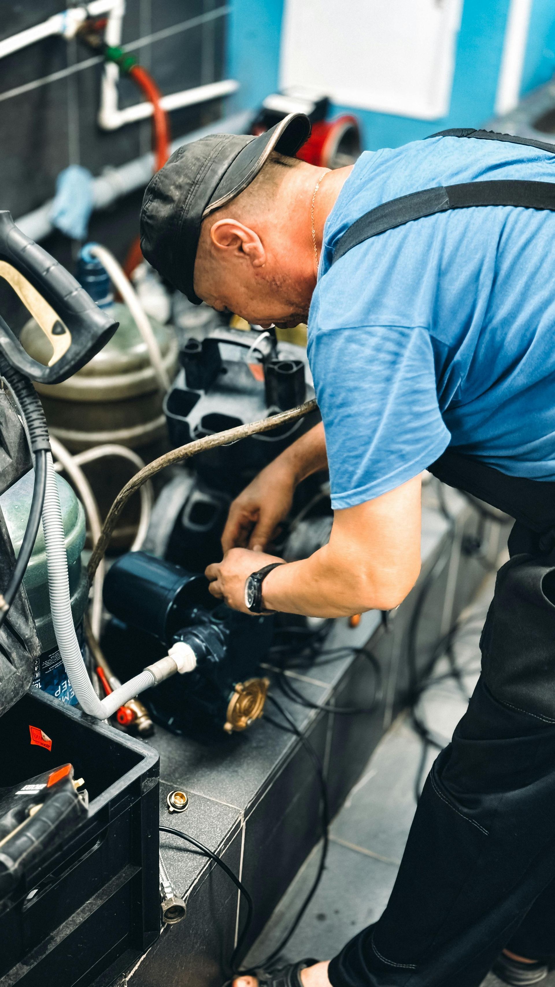 Mechanic in blue shirt works under a vehicle in a garage, using tools near exposed hoses and wiring