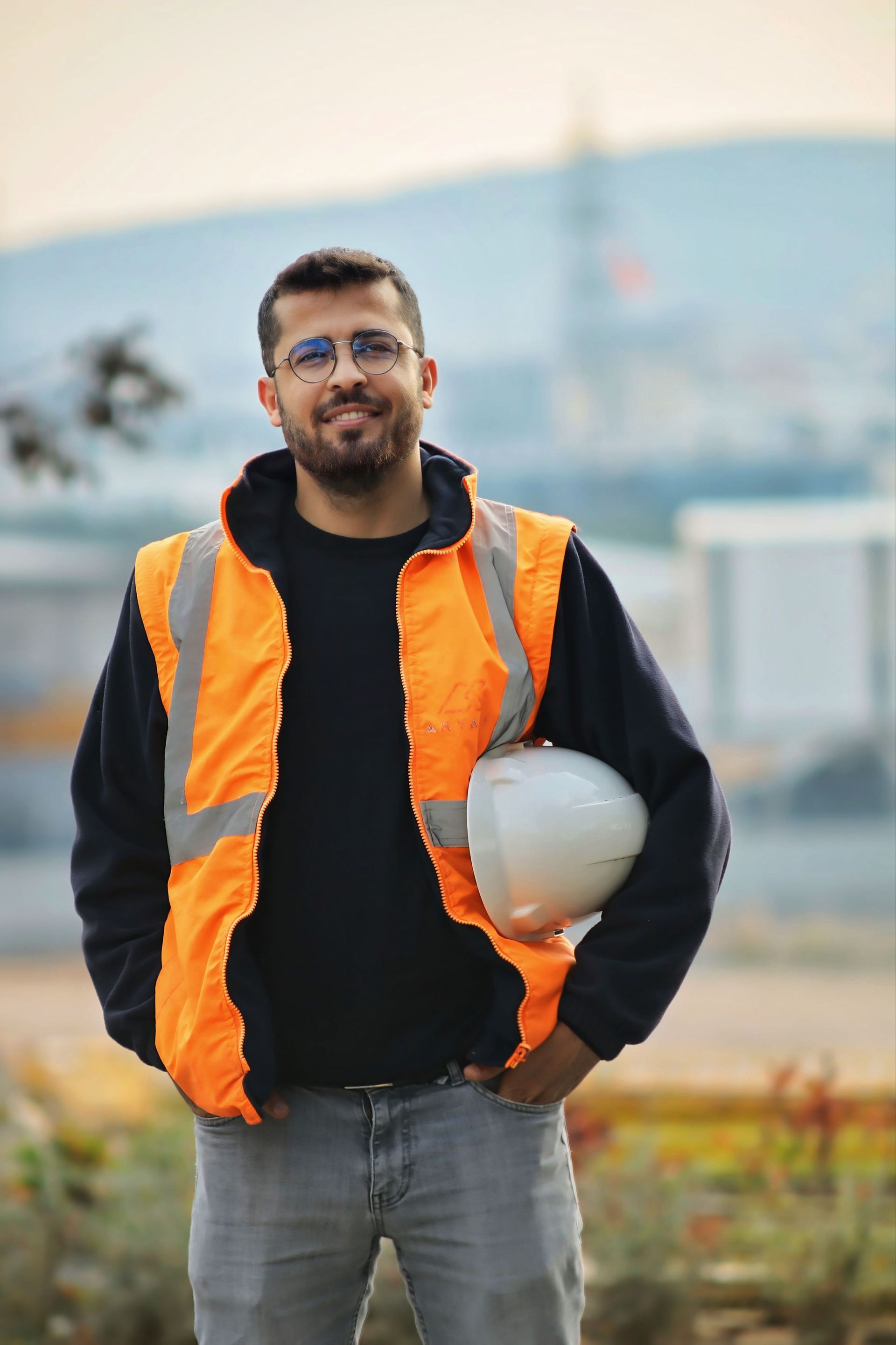Man in orange safety vest holding a white hard hat outdoors at a construction site