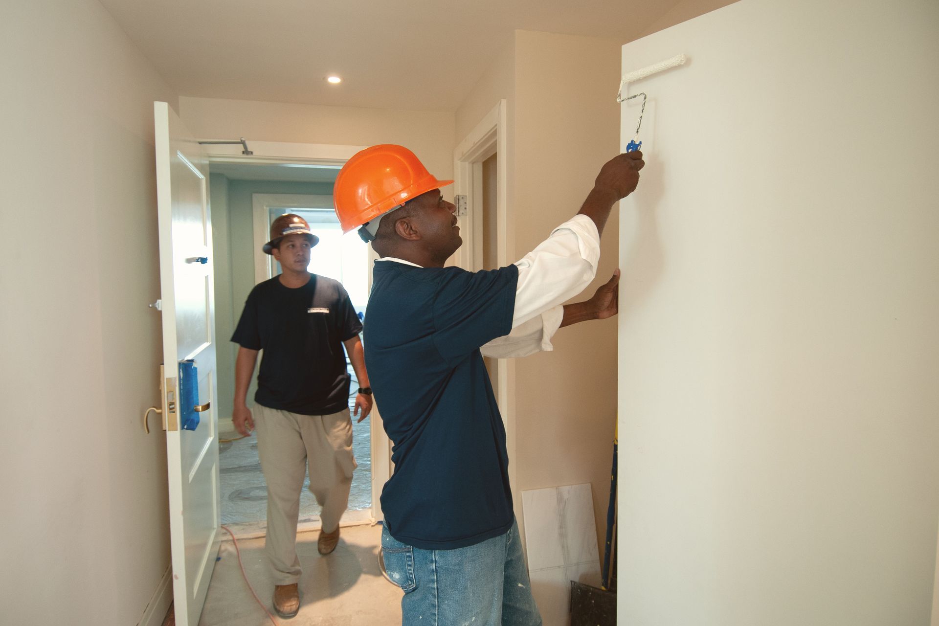 Two workers in a hallway, one in an orange hard hat inspecting a wall with a flashlight.