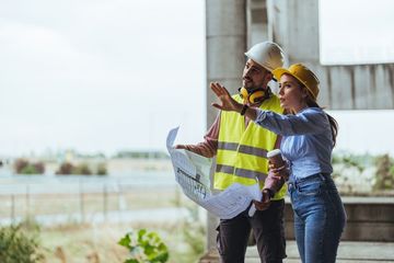 Young woman architect explaining blueprint to supervisor wearing safety vest at construction site.