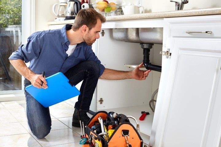 Plumber kneeling under a sink, inspecting pipes with a blue clipboard in a kitchen