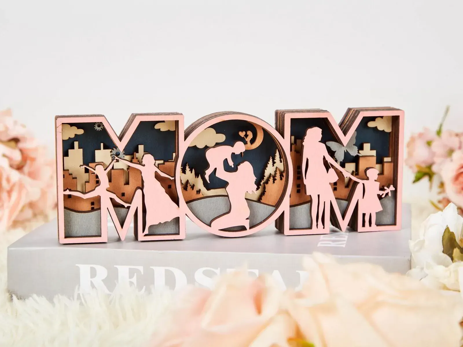 The word mom is cut out of wood and is sitting on top of a book.