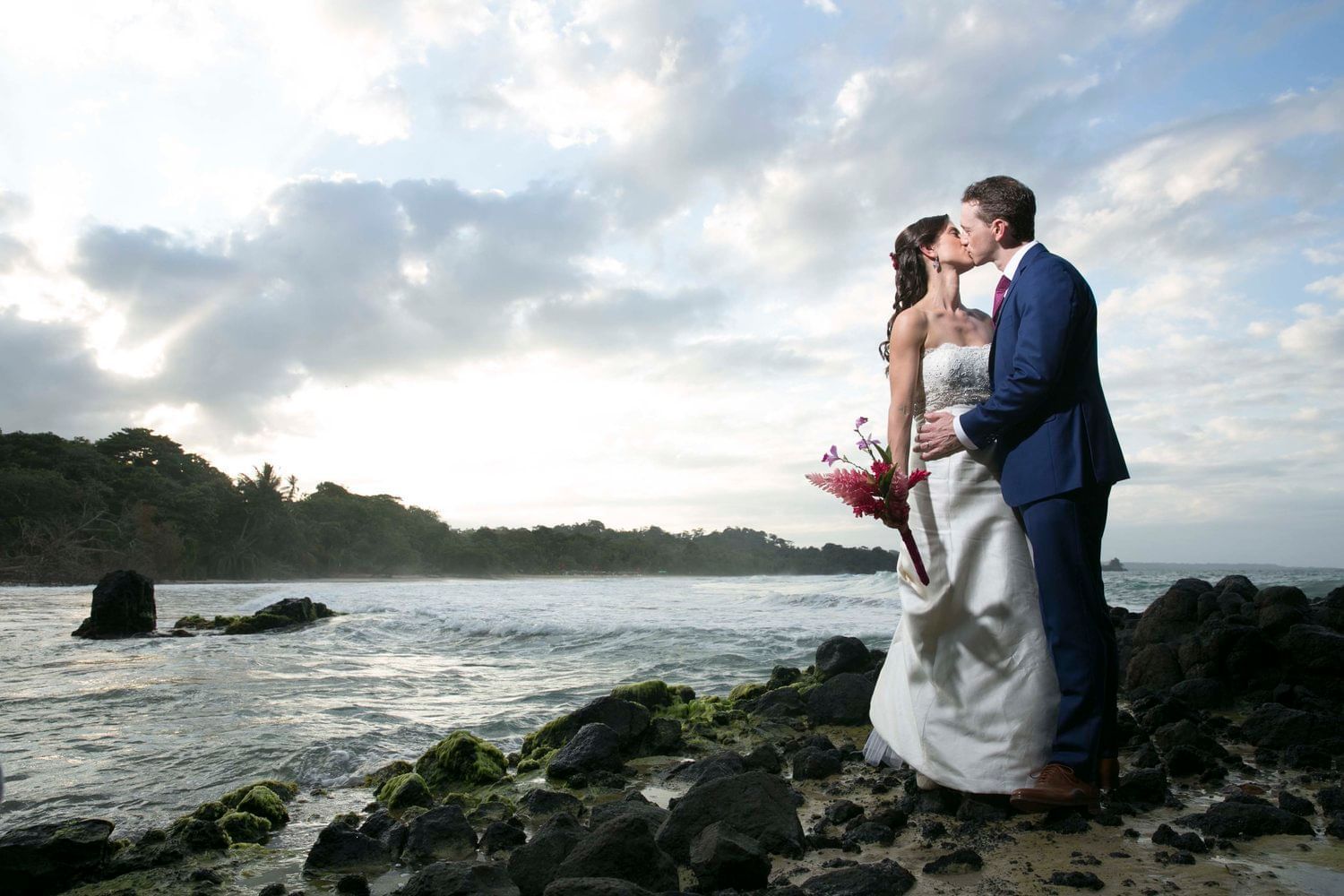 A bride and groom kissing on a rocky beach near the ocean