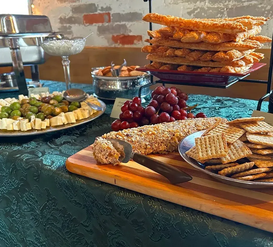 A buffet table features a cheese and olive platter, grapes, a nut-covered cheese log, breadsticks, and crackers.