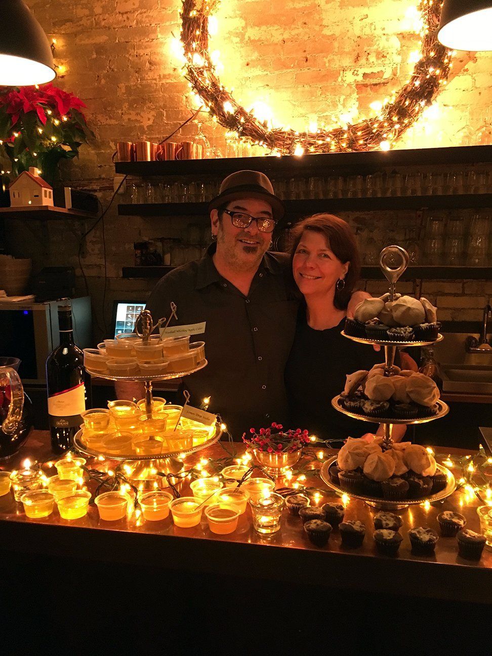 A smiling couple stands behind a bar lit with fairy lights, surrounded by dessert platters and holiday decorations.
