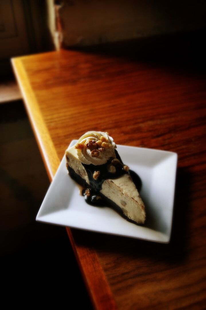A slice of cheesecake topped with whipped cream and chocolate sauce, served on a square white plate on a wooden table.