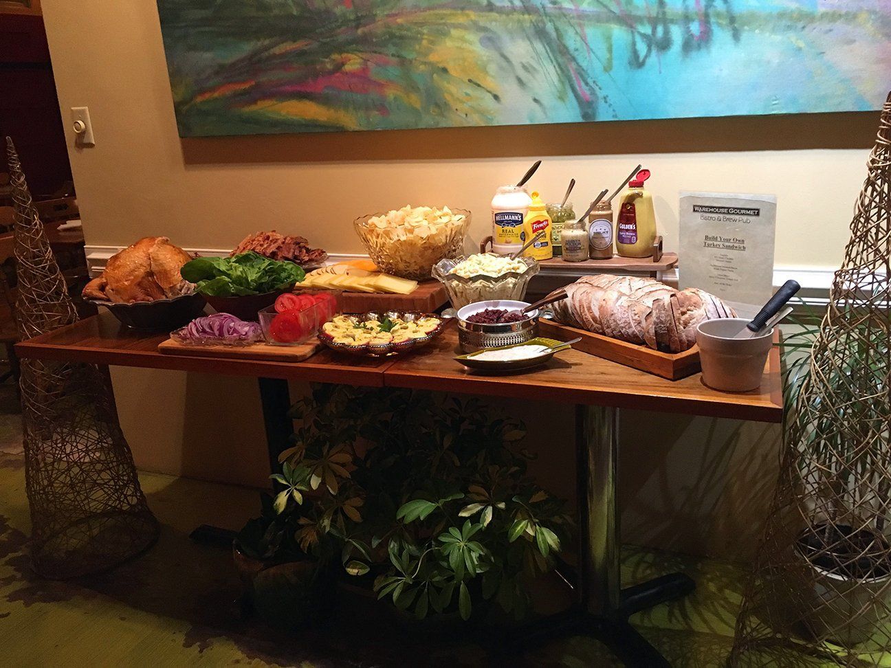 A buffet table featuring platters of deli meats, cheeses, lettuce, tomatoes, onions, condiments, and a bowl of popcorn.