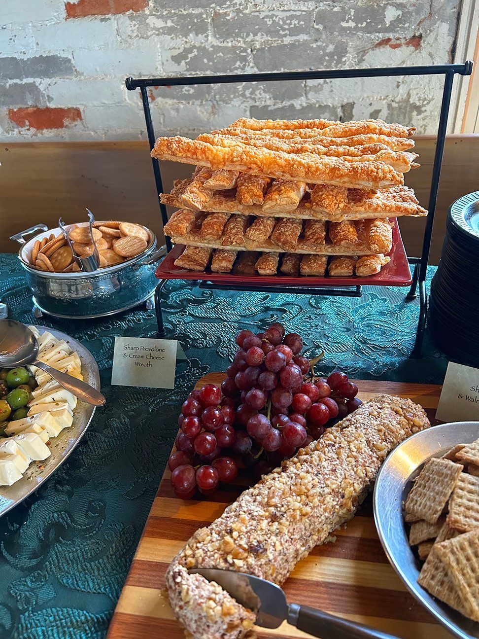A charcuterie spread with stacked breadsticks, grapes, a nut-crusted cheese log, and assorted crackers on a green cloth.