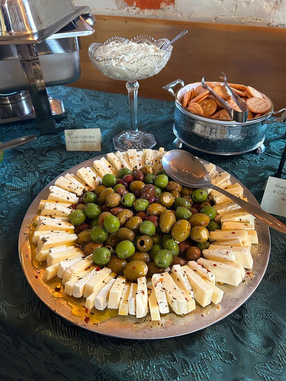 A platter of sliced cheese and green olives sits on a table next to crackers and a bowl of dip on a teal tablecloth.