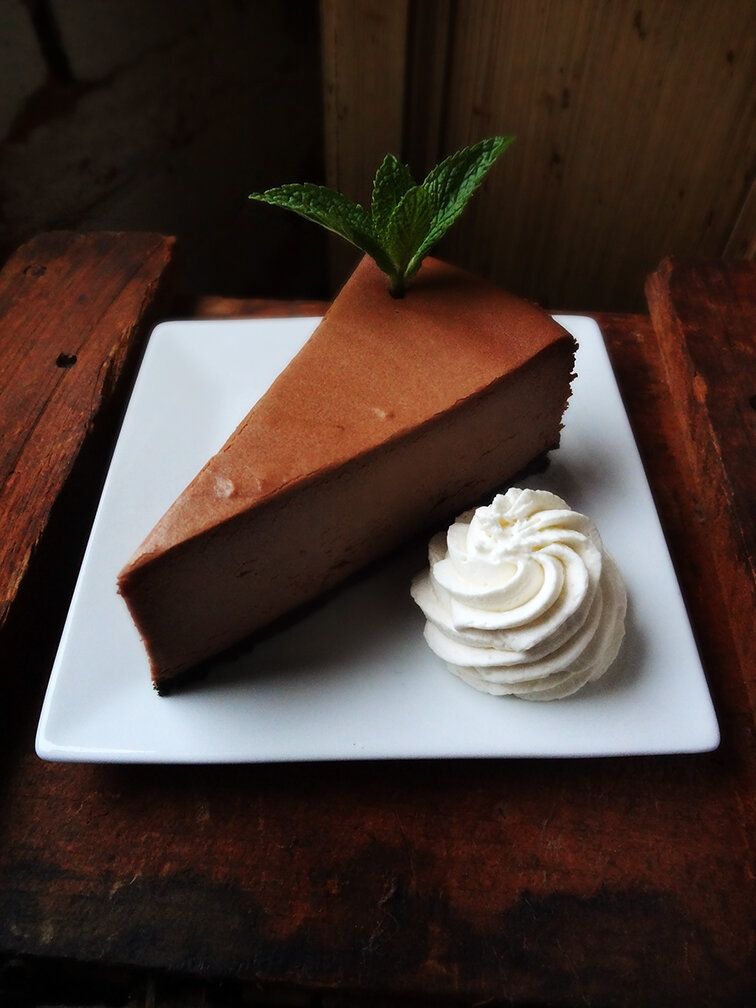 A slice of chocolate cheesecake with a mint garnish and a dollop of whipped cream on a square white plate.
