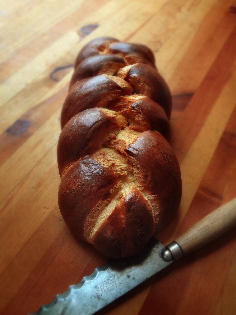 A loaf of golden-brown braided bread rests on a wooden surface next to a serrated bread knife.