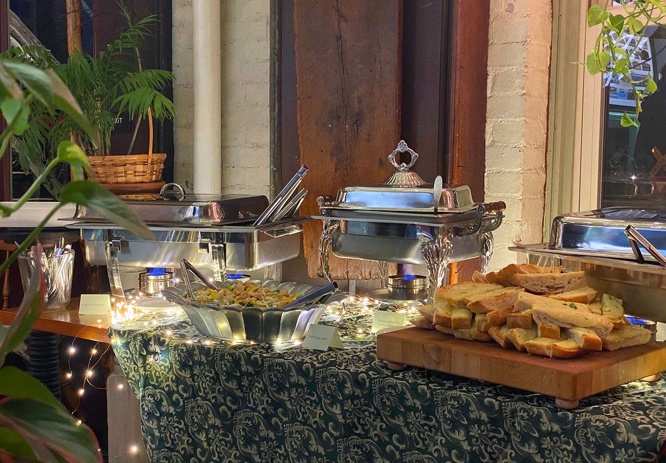 A buffet spread with chafing dishes, a bowl of food, and a wooden board of sliced bread on a patterned tablecloth.