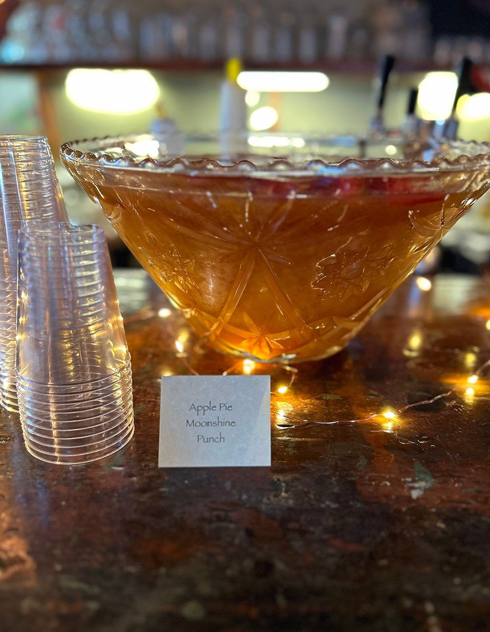 A crystal punch bowl filled with amber liquid sits on a counter next to a stack of plastic cups and a label card.