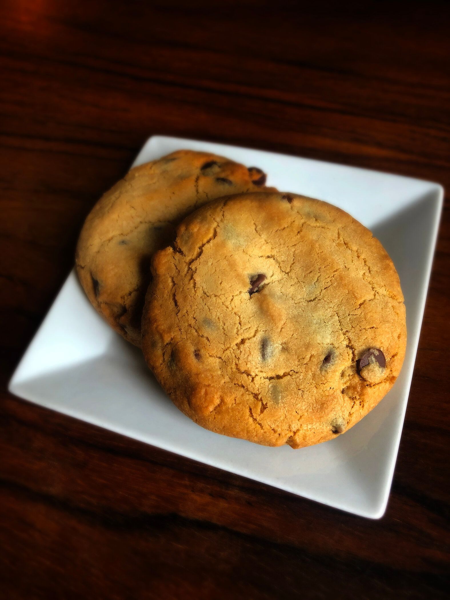 Two chocolate chip cookies resting on a small, white square plate against a dark wooden surface.