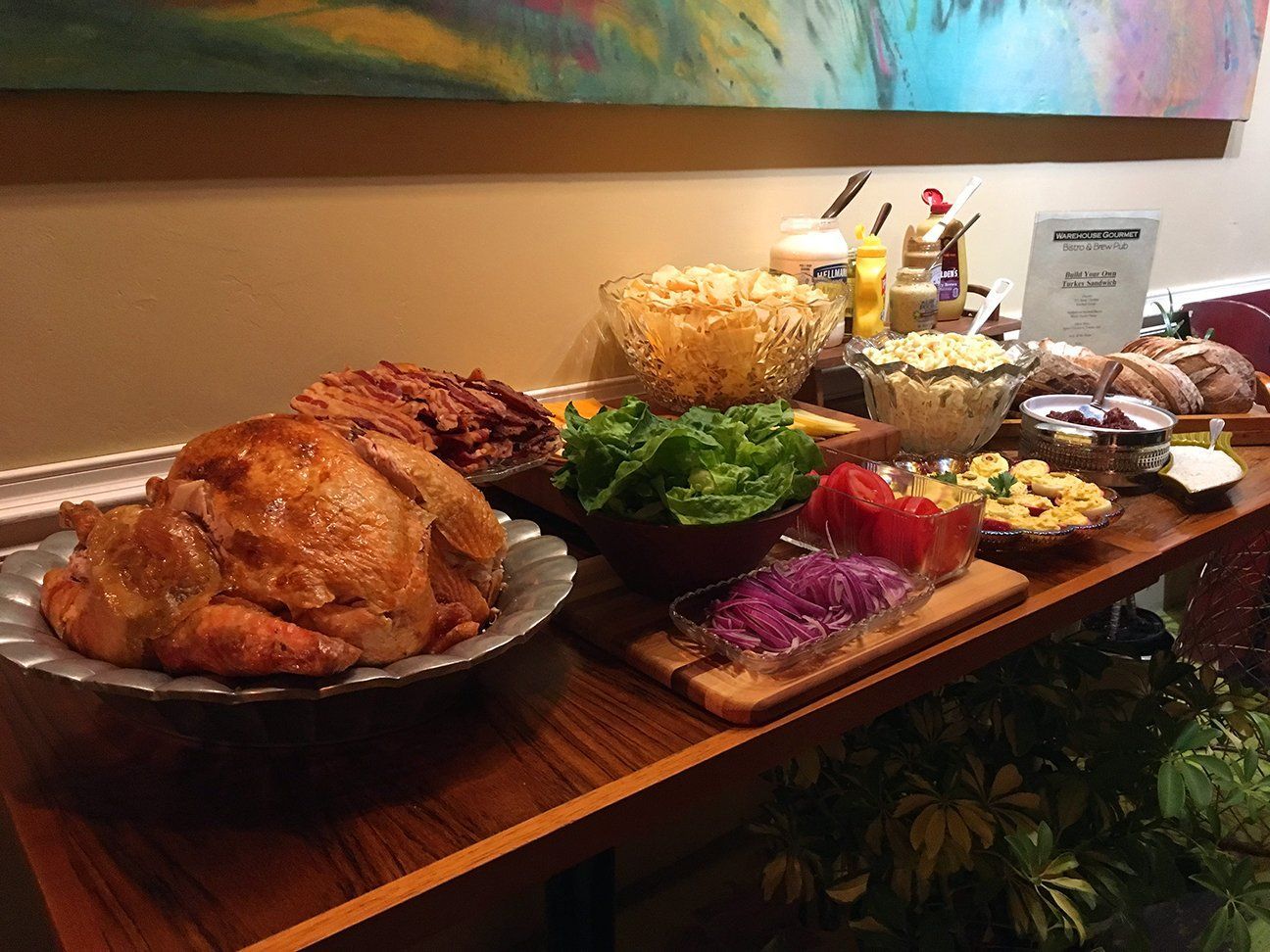 A buffet spread featuring a roasted turkey, sides of lettuce, tomato, red onion, pasta salad, and sliced bread on a table.