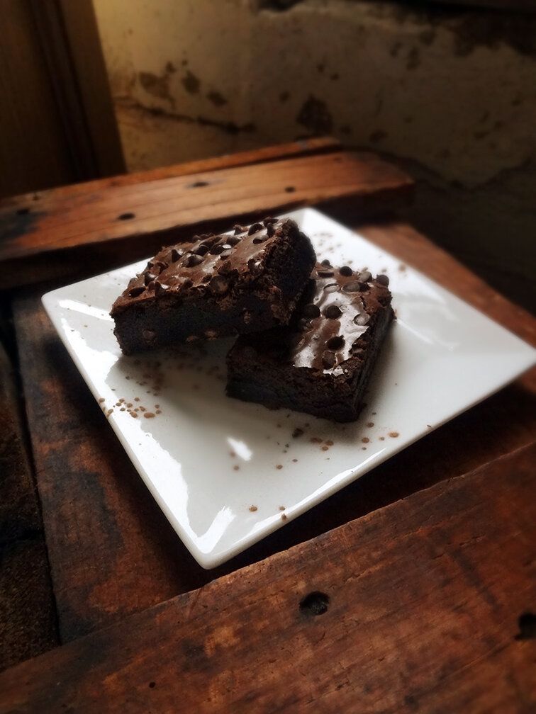 Two chocolate brownies topped with chocolate chips on a white square plate, set on a rustic wooden table.