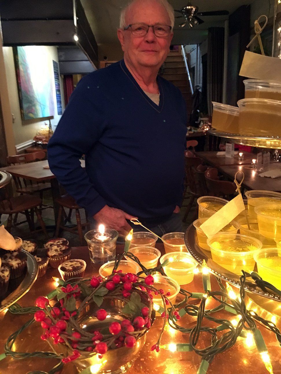 A person in a blue v-neck sweater stands behind a table with festive lighting, small containers of food, and red berries.