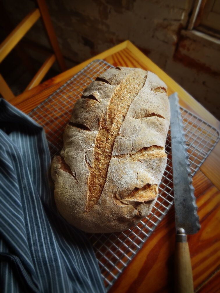 A loaf of artisanal bread with a scored top rests on a wire cooling rack next to a serrated knife on a wooden table.