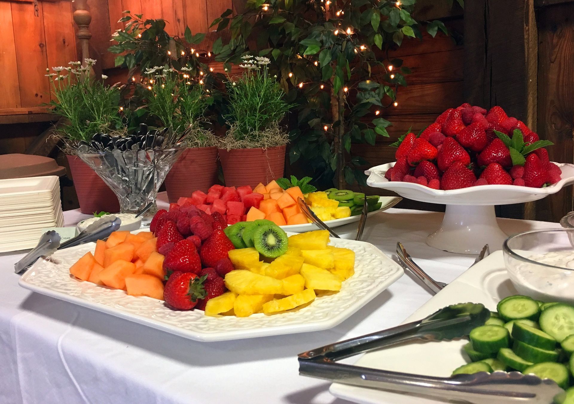 A buffet table features platters of fresh fruit including melon, strawberries, kiwi, and pineapple, alongside cucumbers.