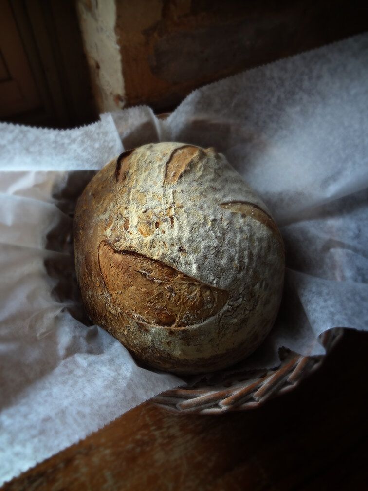 A round, scored loaf of crusty, golden-brown artisan bread rests in a wicker basket lined with white parchment paper.