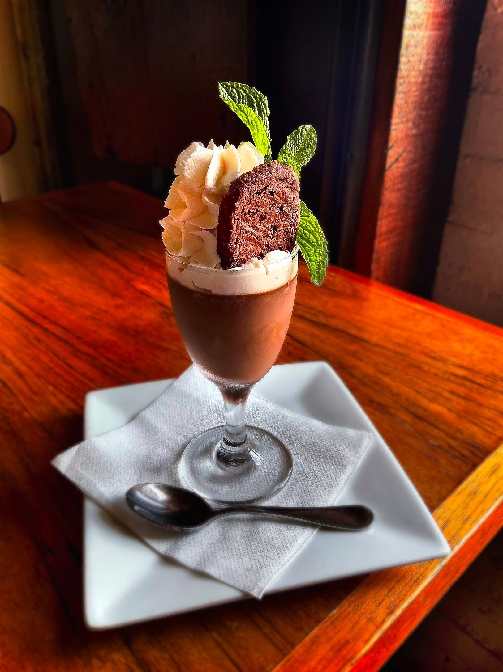 A chocolate mousse dessert in a stemmed glass, topped with whipped cream, a chocolate crisp, and mint, on a square plate.