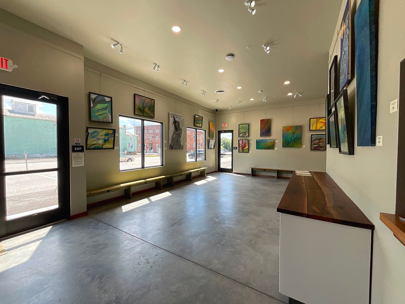 A brightly lit gallery space with paintings displayed on beige walls, concrete flooring, and a long wooden counter.