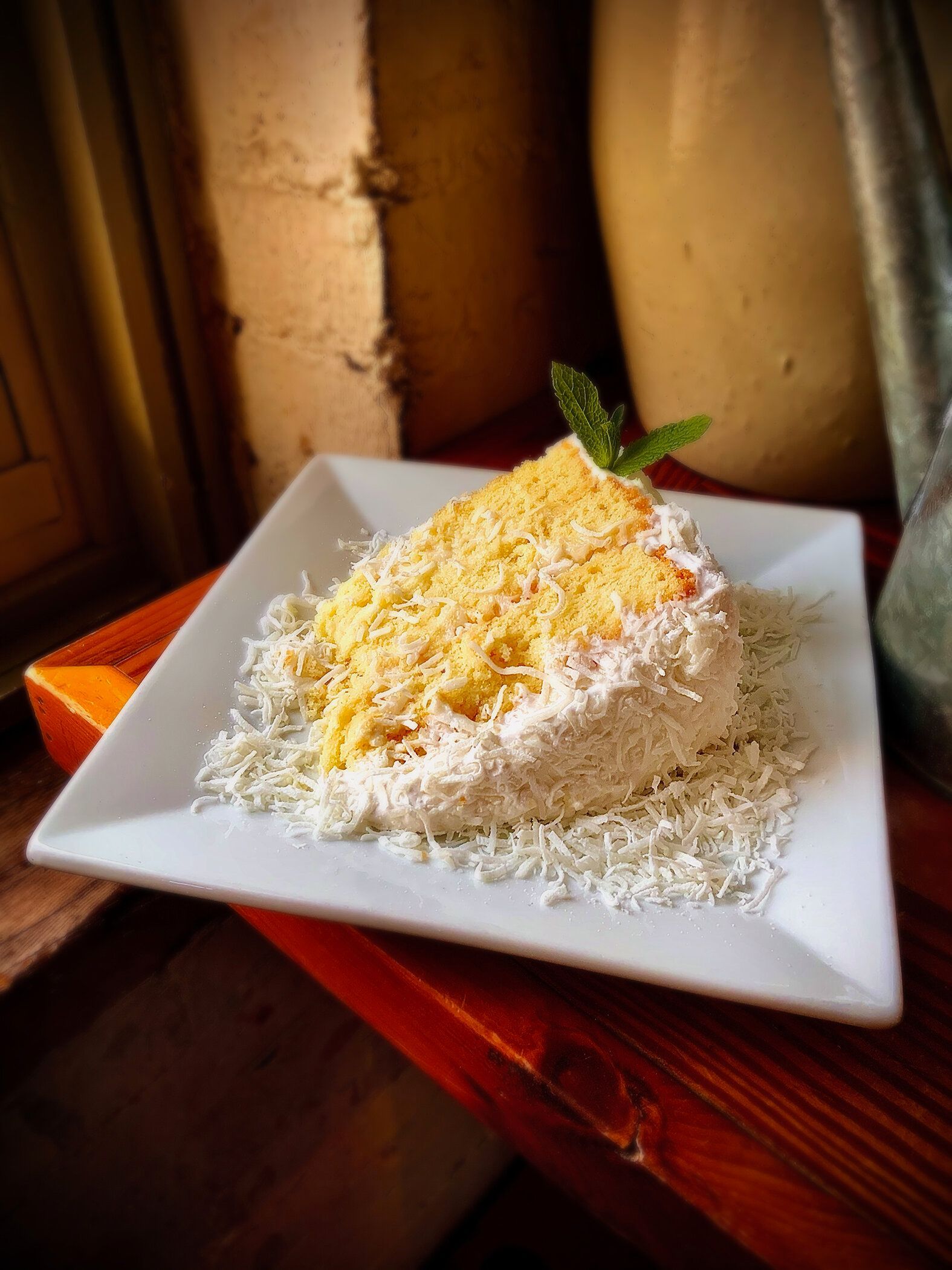 A slice of yellow cake covered in white coconut shavings on a square white plate, garnished with a sprig of mint.