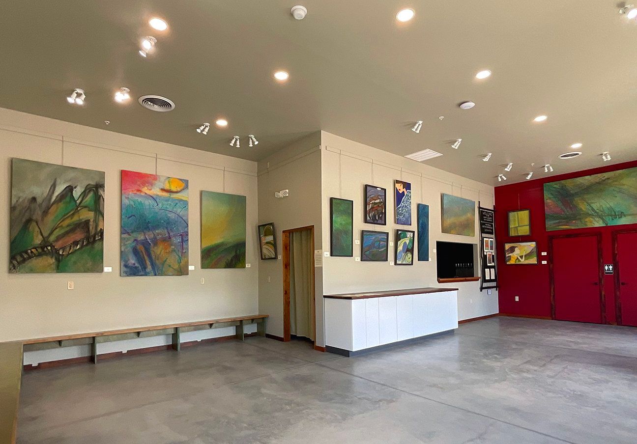 An art gallery interior with paintings on light beige walls and a red accent wall, featuring concrete floors and lighting.