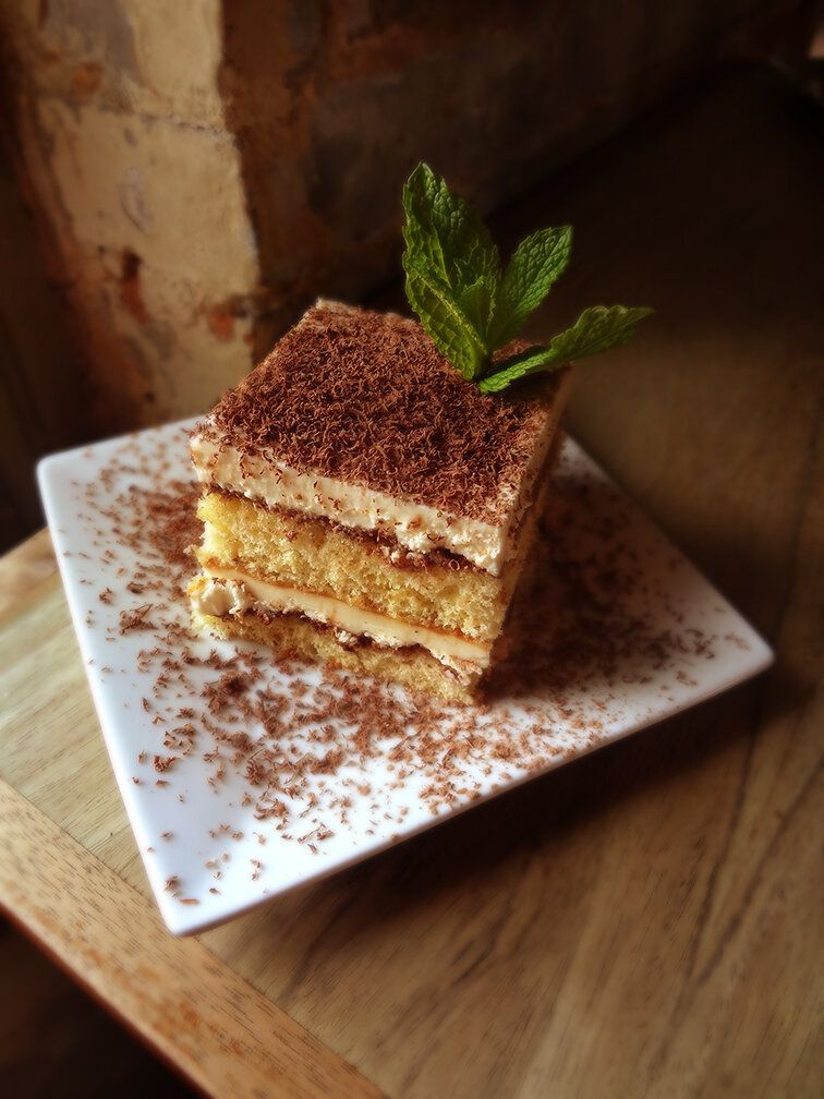 A square piece of tiramisu topped with mint, dusted with cocoa on a white plate over a wooden table.