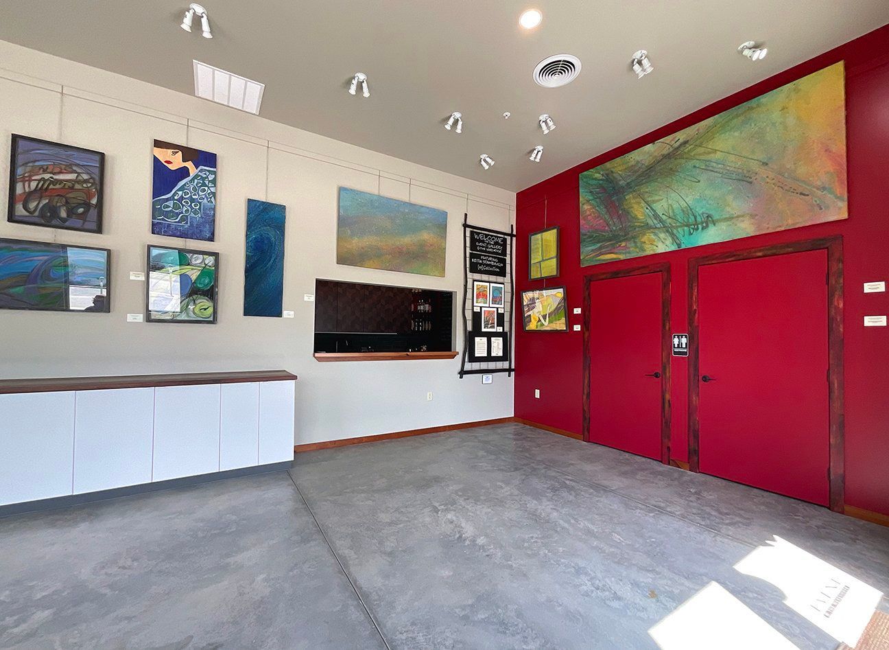 An art gallery interior with paintings on white and red walls, a concrete floor, and a white storage unit.