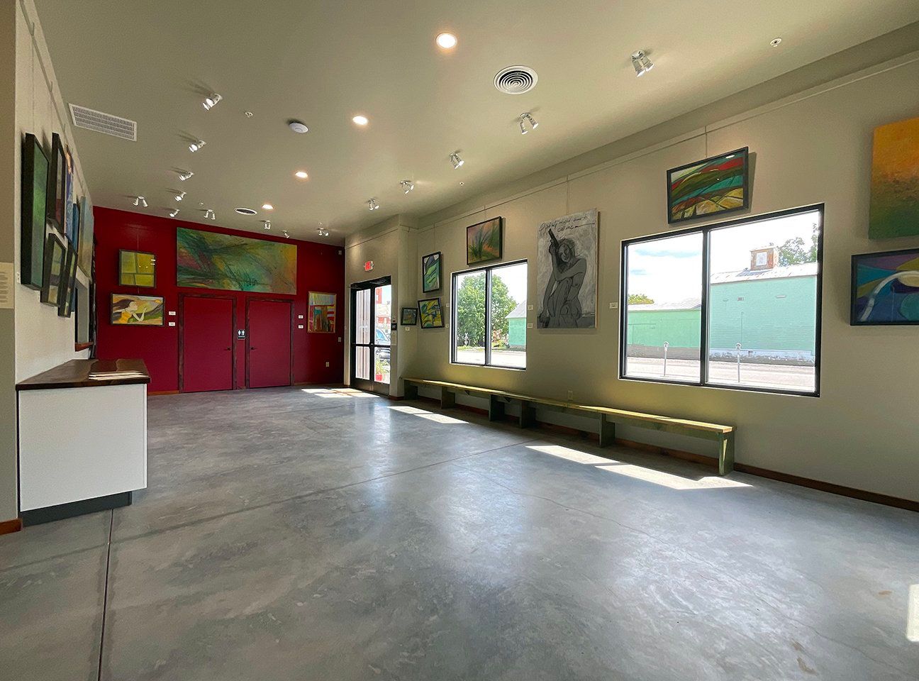 An art gallery with concrete floors, a red accent wall, large windows, and framed paintings displayed on white walls.