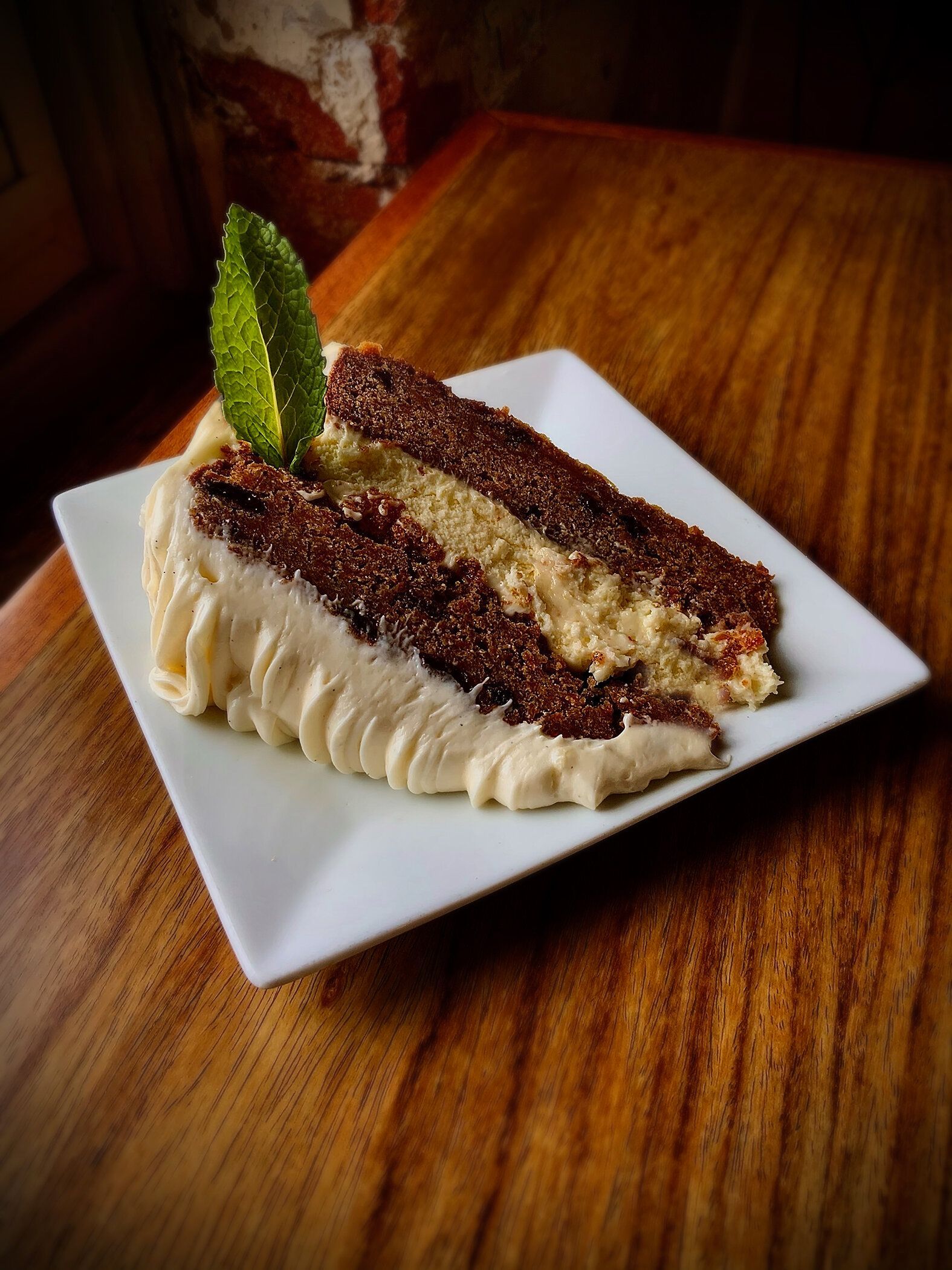 A slice of layered chocolate cake with creamy frosting and a mint leaf garnish, served on a white plate on a wood table.