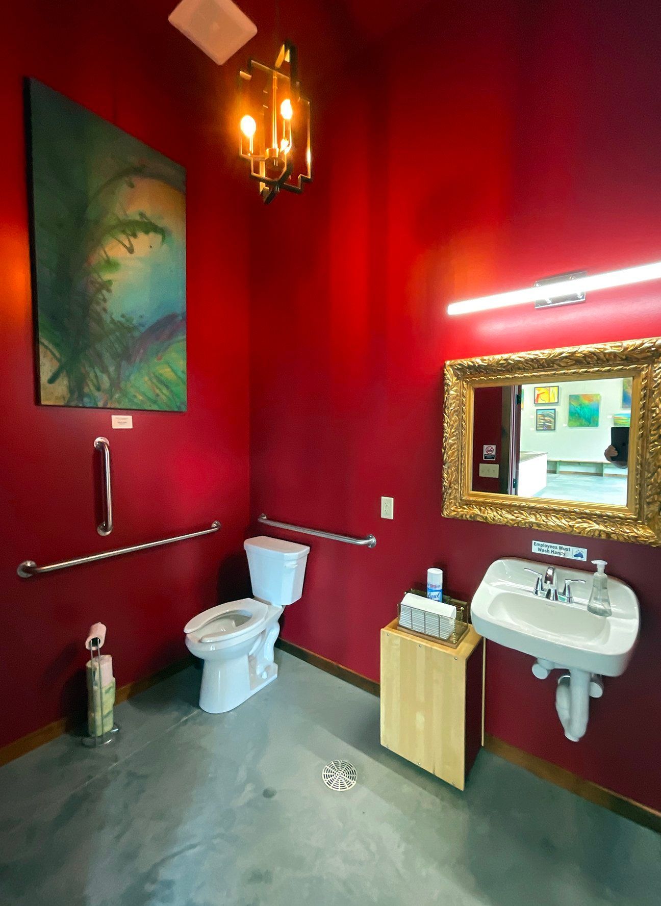 An accessible restroom with deep red walls, a toilet, a wall-mounted sink with an ornate gold mirror, and wall grab bars.