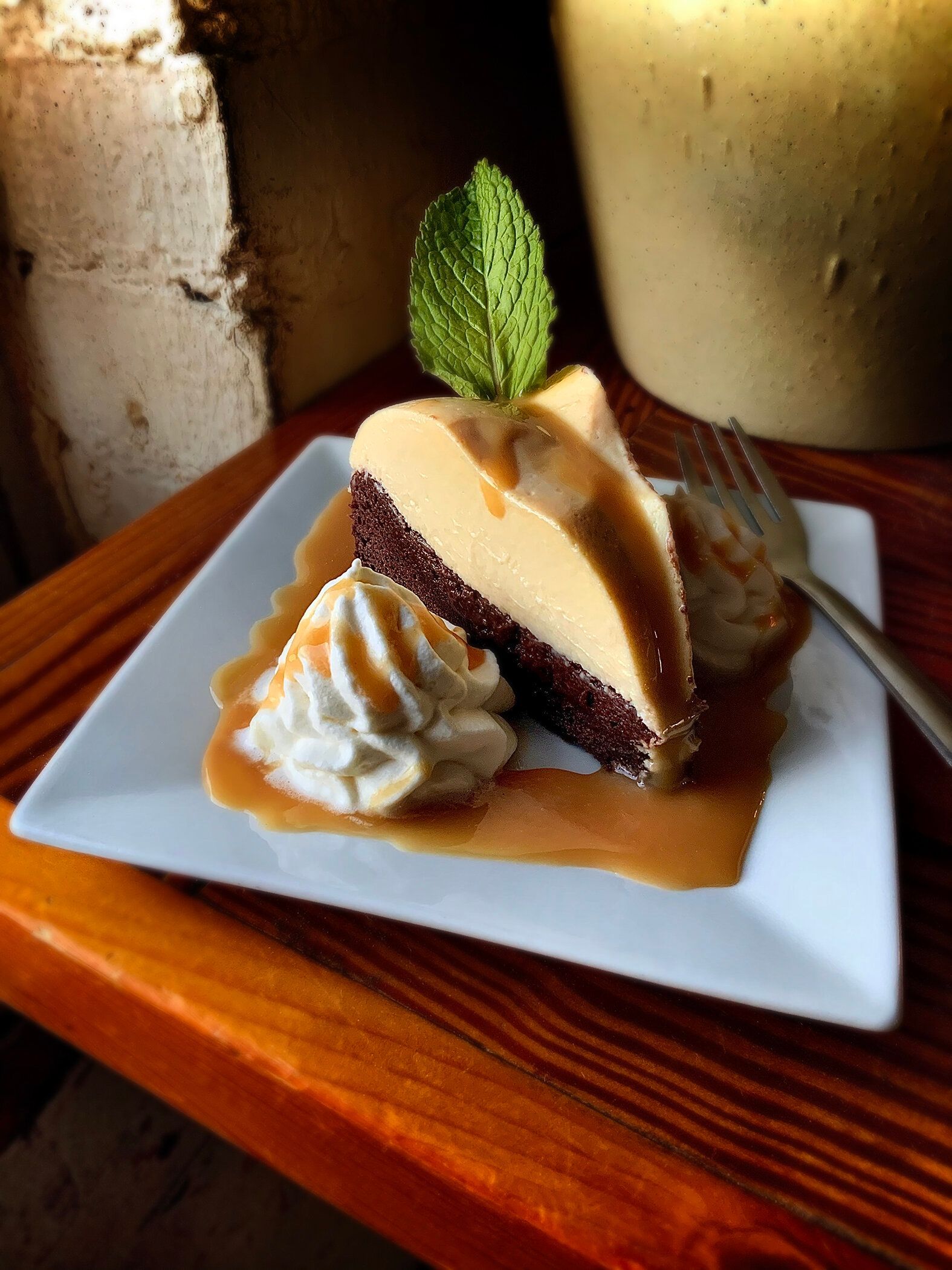 A slice of chocolate cake topped with custard, caramel sauce, a dollop of whipped cream, and a fresh mint leaf on a plate.